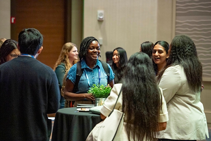 Group of attendees standing around a tall round table during a networking session. A small potted plant and notebooks are on the table.
