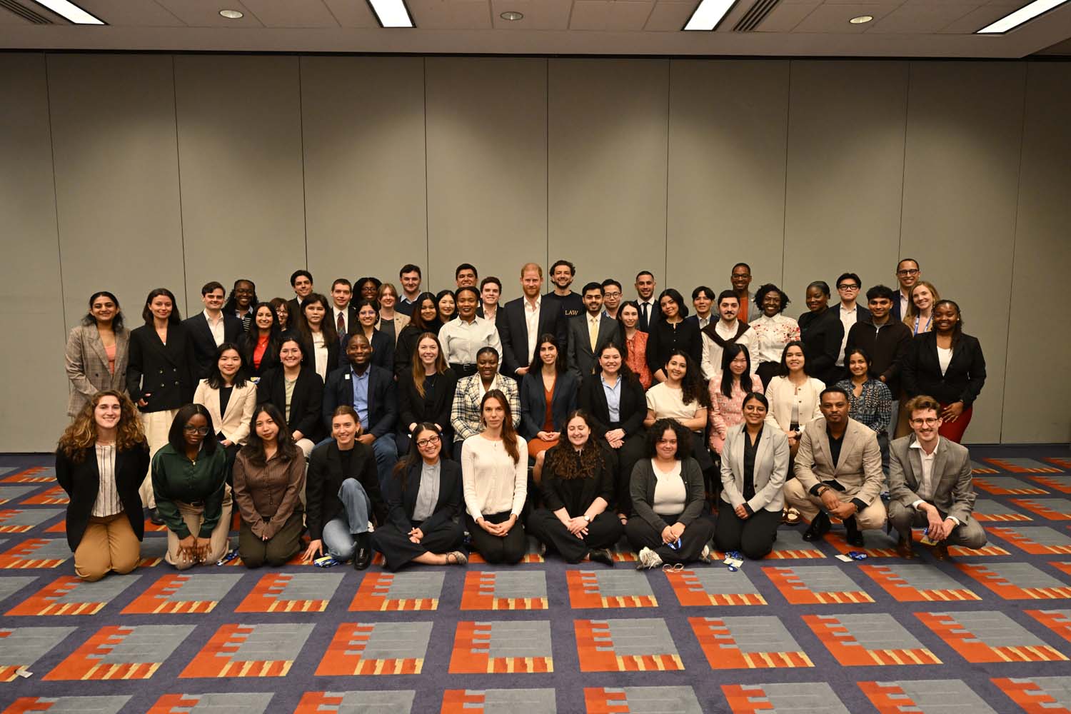 A large group of conference participants poses for a formal photo in multiple rows inside a meeting room, all facing the camera against a plain wall backdrop.