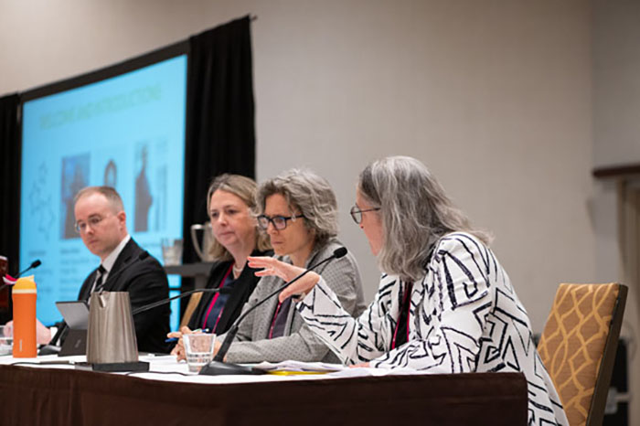 Panel discussion with four speakers seated at a table, microphones and water glasses in front. A large screen behind displays presentation slides.