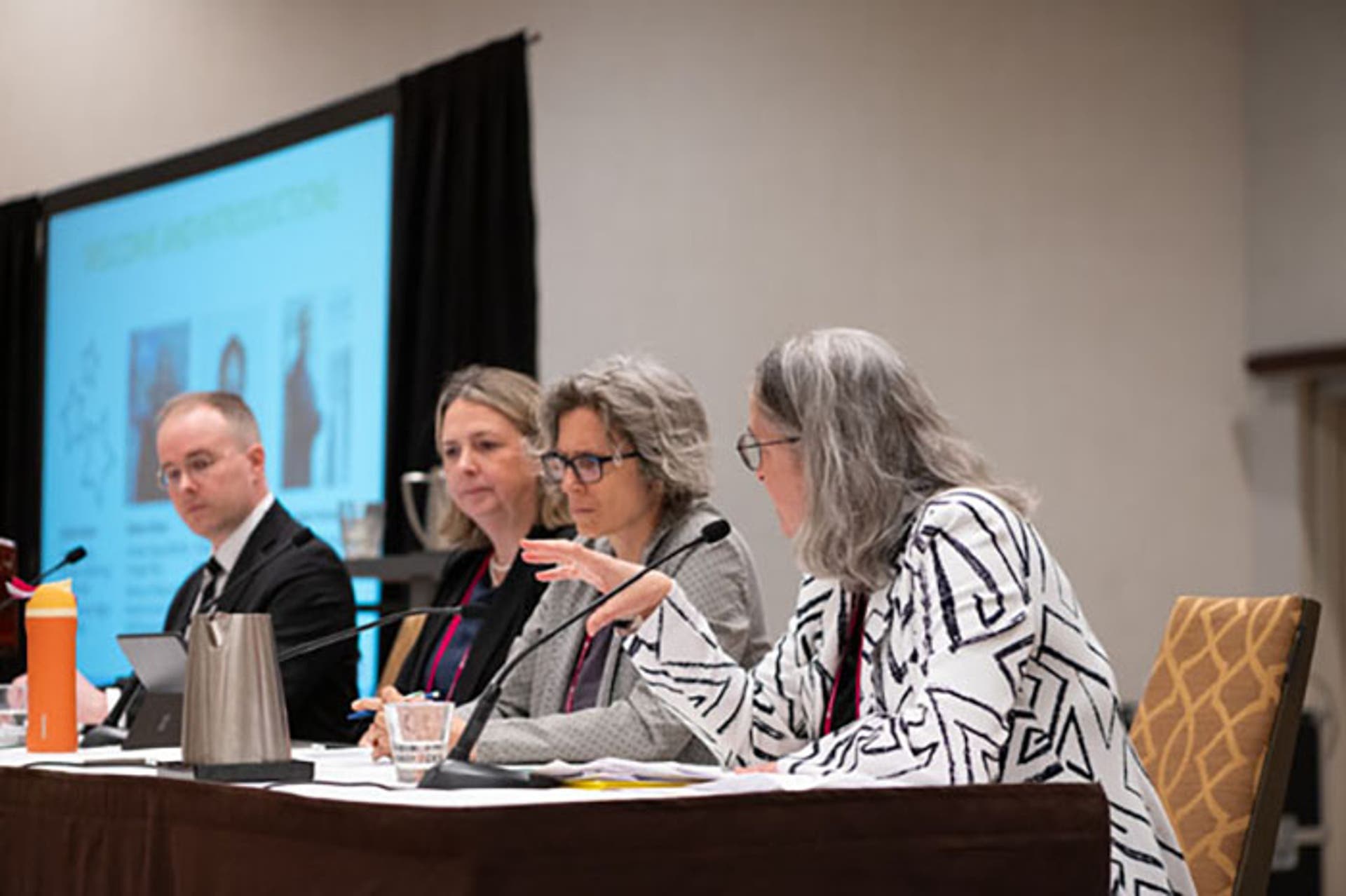 Panel discussion with four speakers seated at a table, microphones and water glasses in front. A large screen behind displays presentation slides.