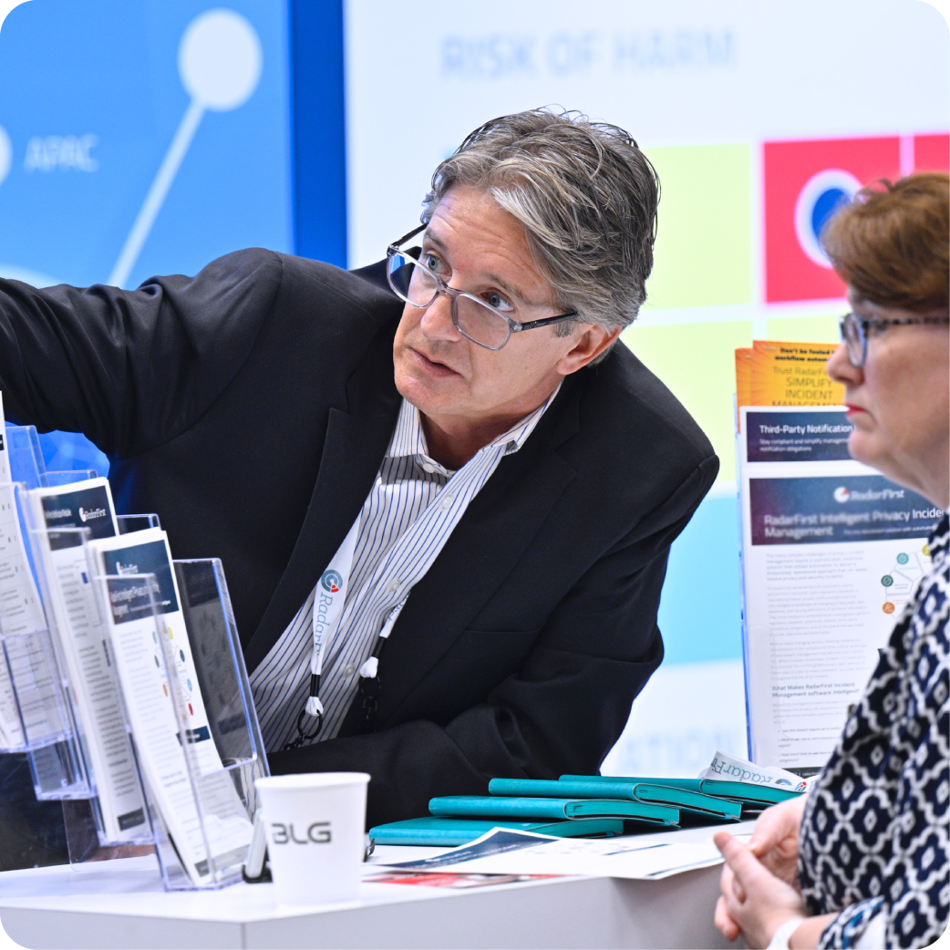 A man in a blue suit jacket and glasses is pointing to something outside the picture. He is talking with another woman in glasses.