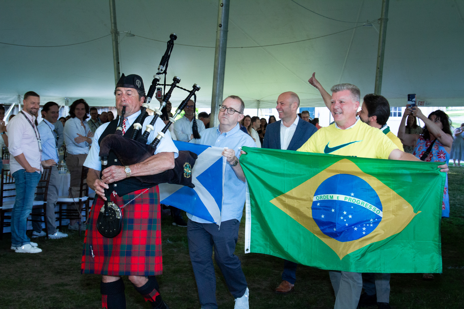 A bagpiper in traditional Scottish dress leads a procession through a large event tent while attendees carry the flags of Scotland and Brazil, creating a festive international atmosphere as onlookers gather along the sides to watch.