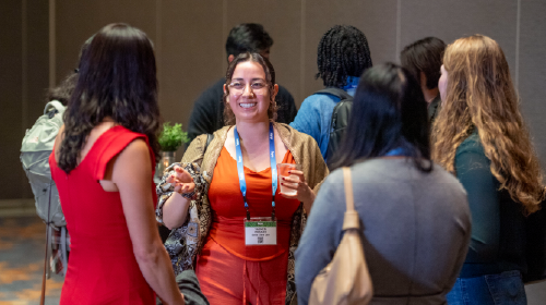A young woman with glasses and wearing an orange dress is talking to four other people standing around her. She is smiling and excited.