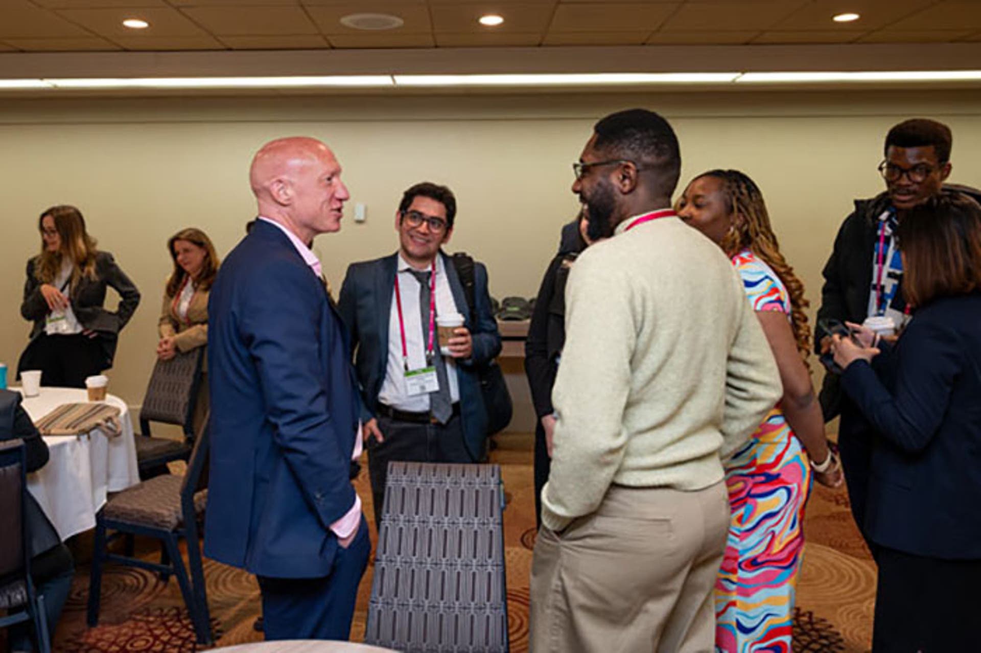 Small group of attendees standing and talking in a meeting room, holding coffee cups and wearing conference lanyards.