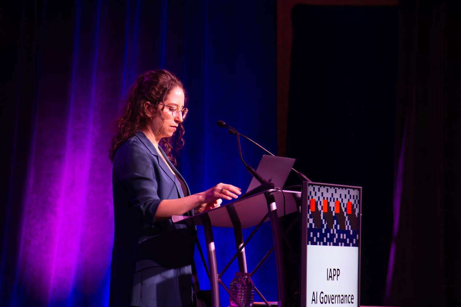 A speaker stands at a podium on a stage washed in purple and blue light, referencing notes on a laptop while addressing the audience at an IAPP AI Governance event.