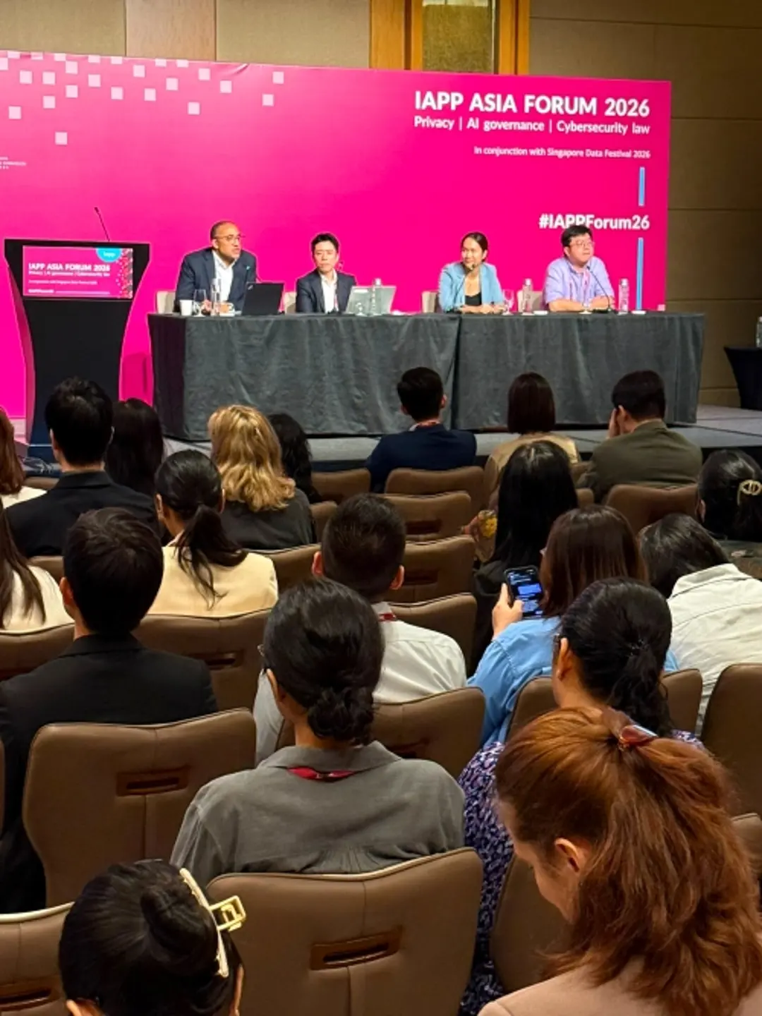 A panel discussion takes place on a stage at the IAPP Asia Forum 2026 conference, with four speakers seated behind a table in front of a large pink event backdrop. Audience members are seated in rows facing the stage, and conference branding for privacy, AI governance, cybersecurity, and law is visible behind the speakers.