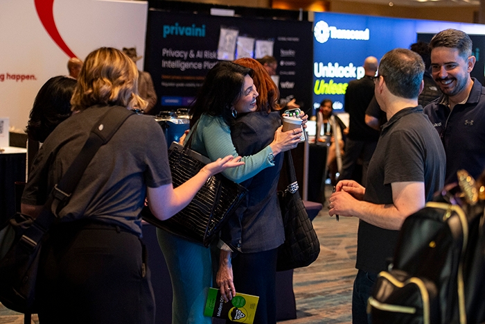 Attendees greeting each other in an exhibit hall, with one person holding a coffee cup and another carrying a tote bag. Event booths and banners are visible in the background.