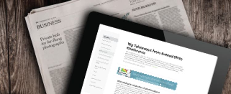 An iPad displaying IAPP news content rests at an angle on a rustic wood surface, partially covering a folded newspaper showing the Business section.