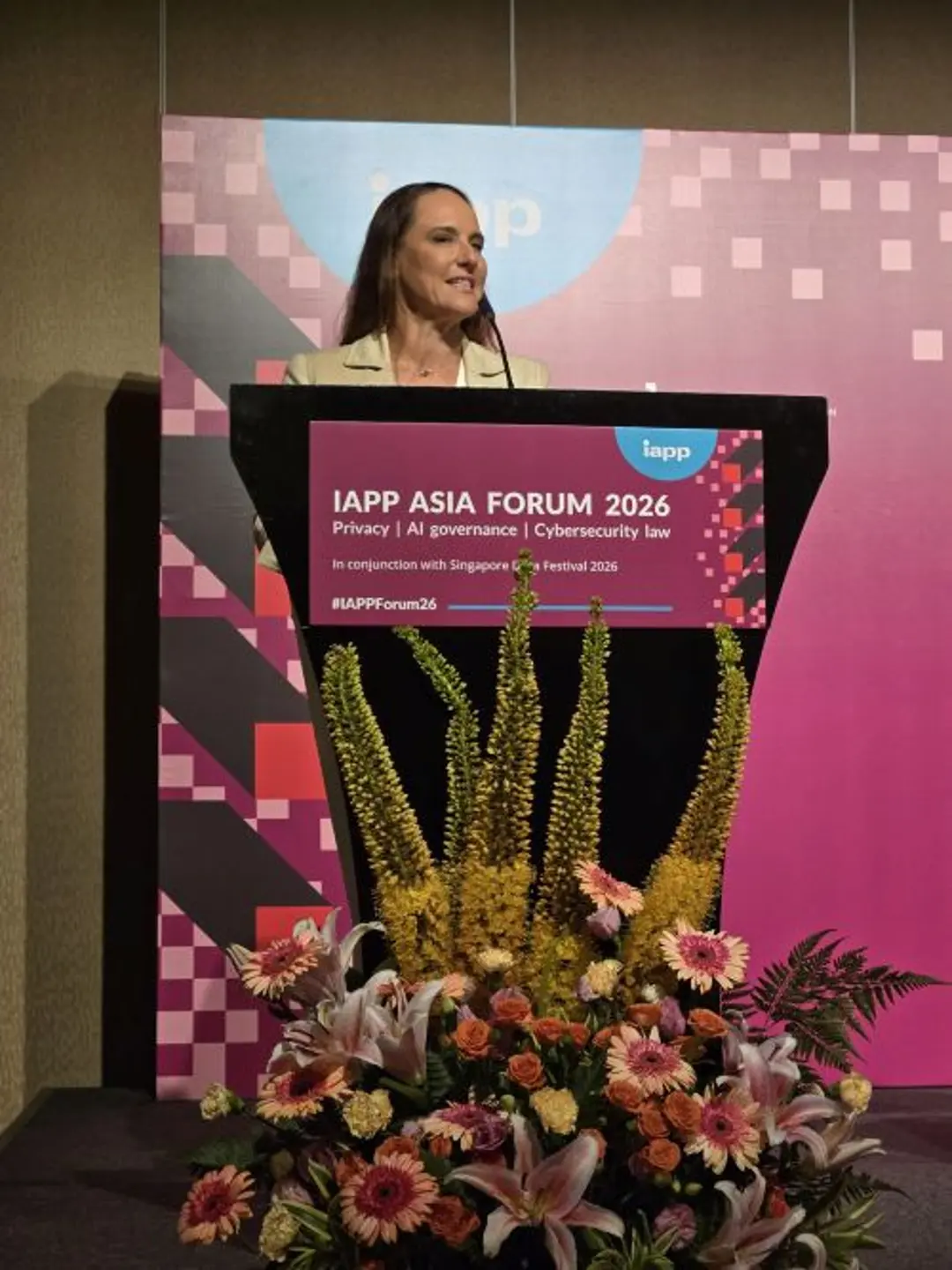 A speaker stands behind a podium at the IAPP Asia Forum 2026, addressing attendees from the stage. A large floral arrangement decorates the front of the podium, and the event branding for privacy, AI governance, cybersecurity, and law is displayed on a pink conference backdrop behind the speaker.