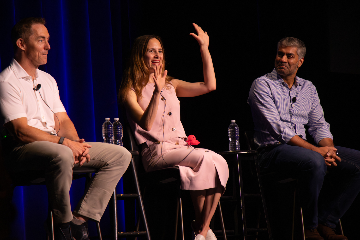 Three speakers sit on stage during a panel discussion, framed by deep blue curtains, as the central panelist gestures with raised hands while speaking and the other panelists listen from either side with microphones and water bottles nearby.