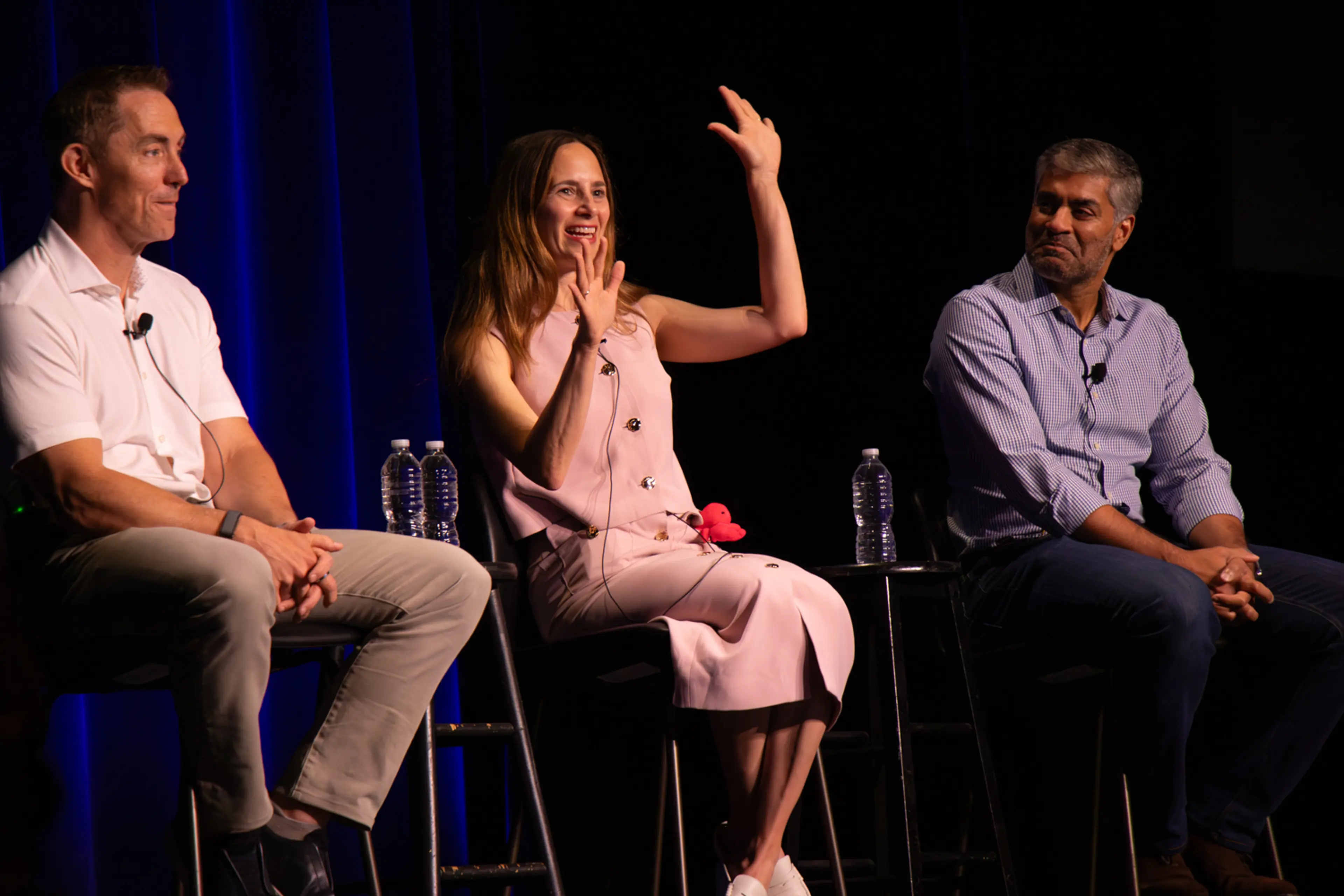 Three speakers sit on stage during a panel discussion, framed by deep blue curtains, as the central panelist gestures with raised hands while speaking and the other panelists listen from either side with microphones and water bottles nearby.