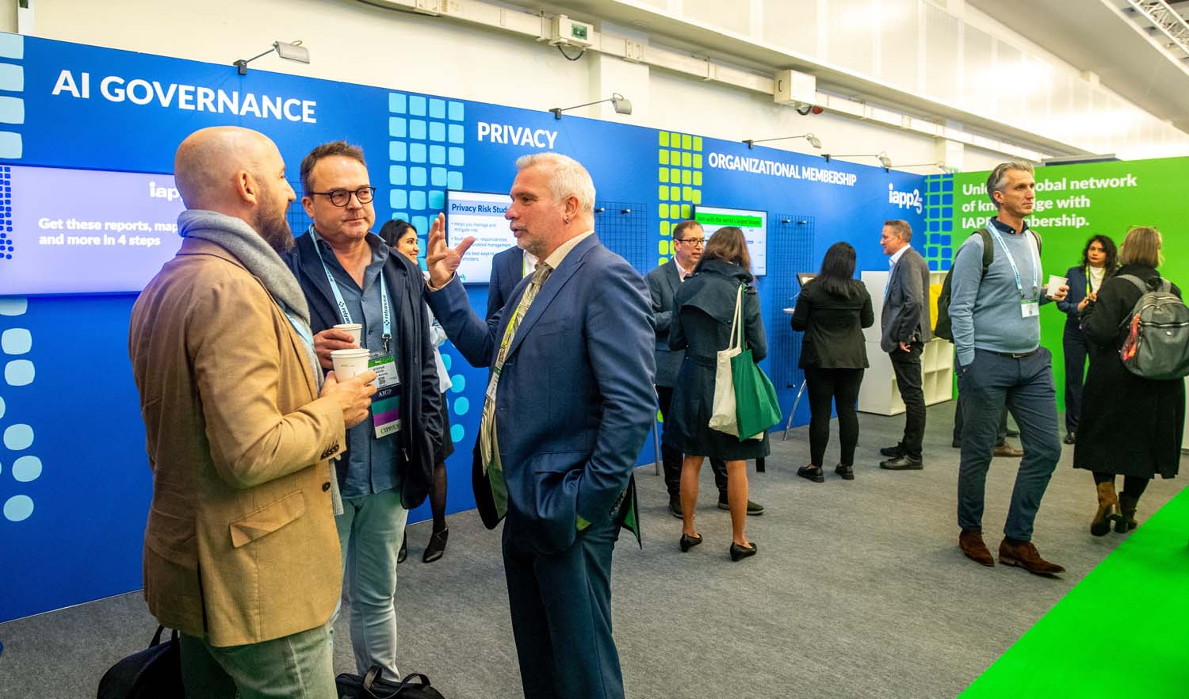 Conference exhibit area with attendees standing and talking in front of a bright blue display wall featuring sections labeled “AI Governance,” “Privacy,” and “Organizational Membership.” A green banner promoting IAPP membership is visible on the right.