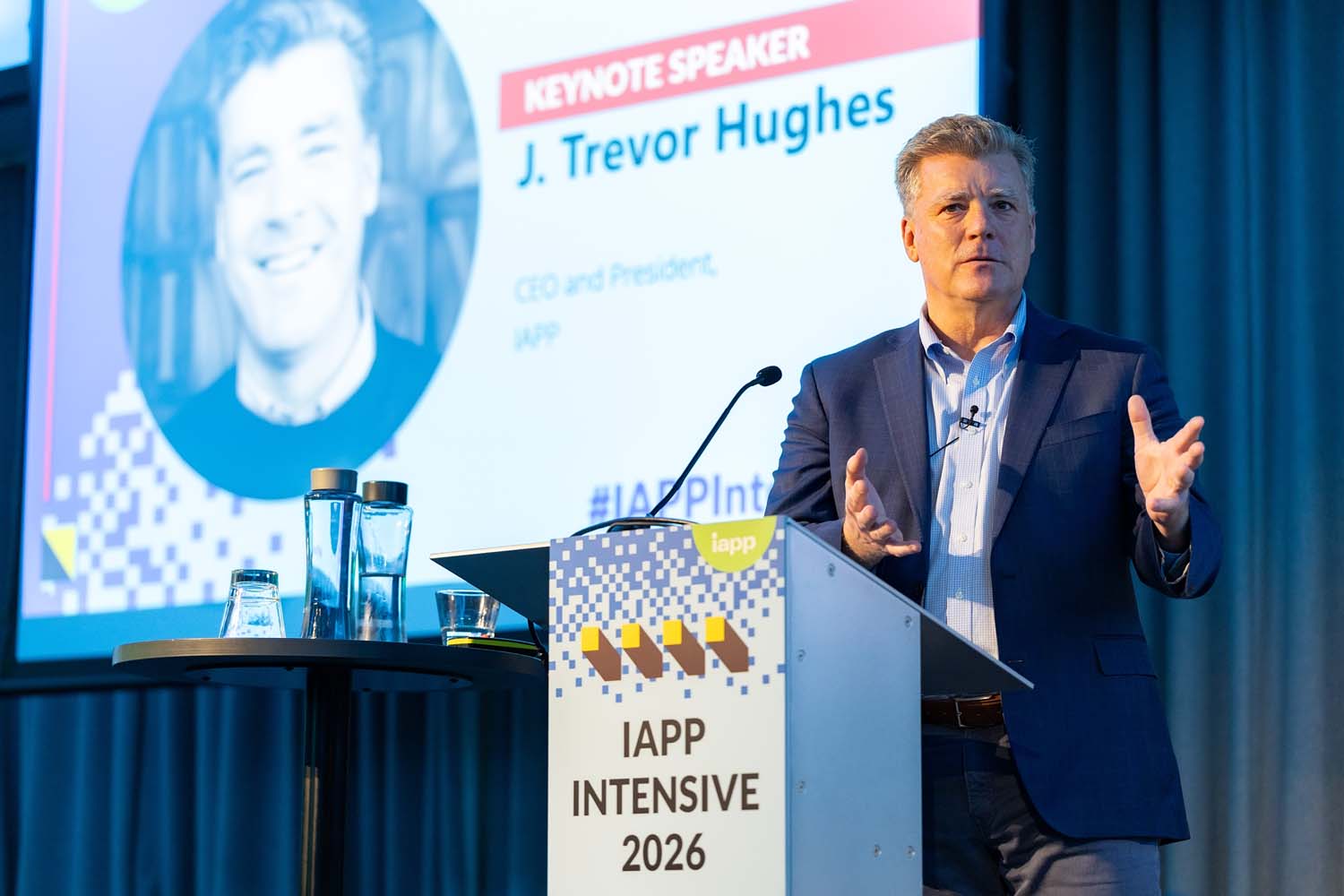 A speaker presents from a podium labeled IAPP Intensive 2026 with a large screen behind the stage showing keynote speaker graphics. Conference materials and water glasses sit on a side table.