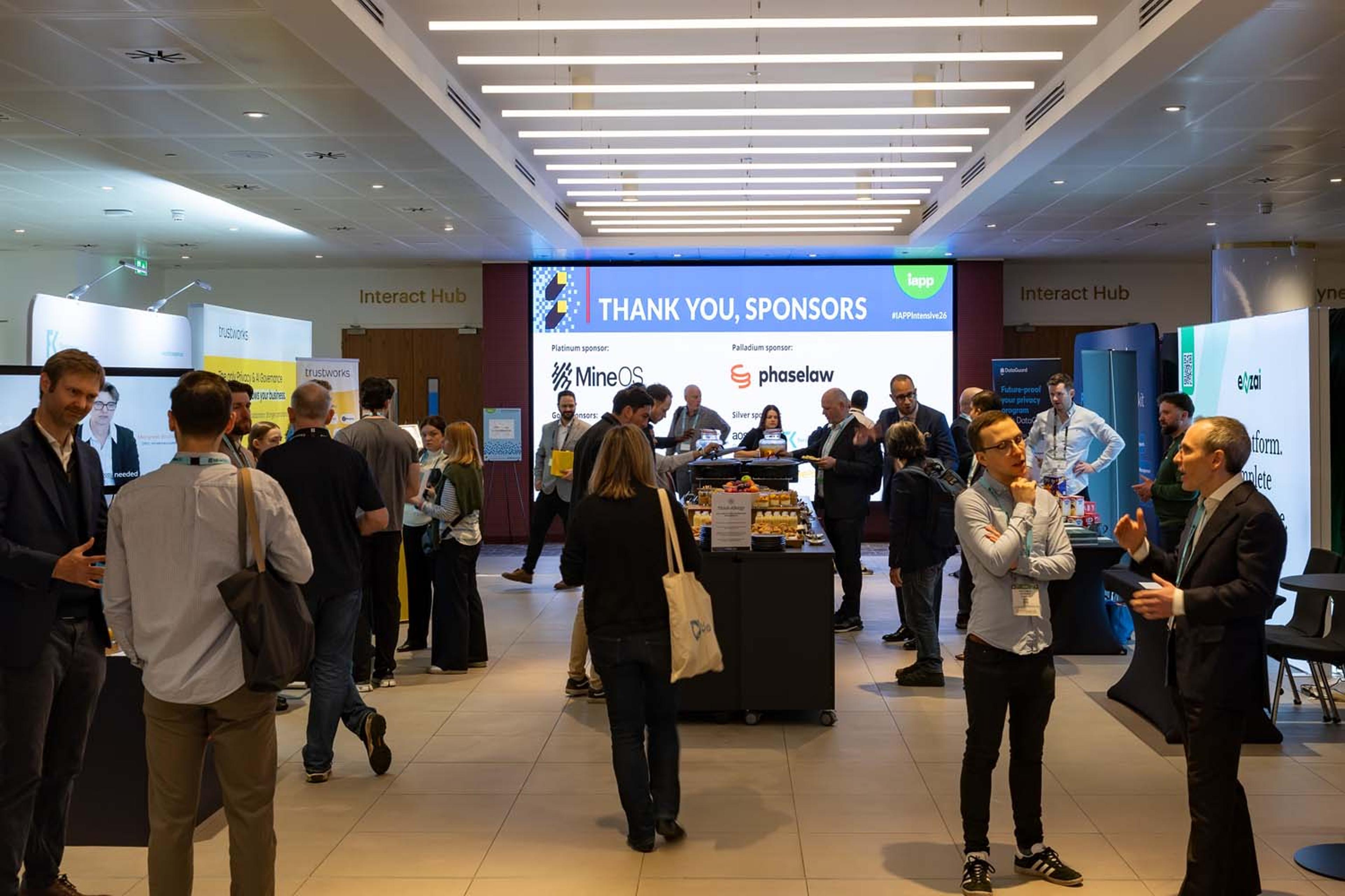 Attendees network and converse in a conference expo area with sponsor booths and a large Thank You Sponsors sign visible in the background. People stand in small groups throughout a bright, open lobby space.