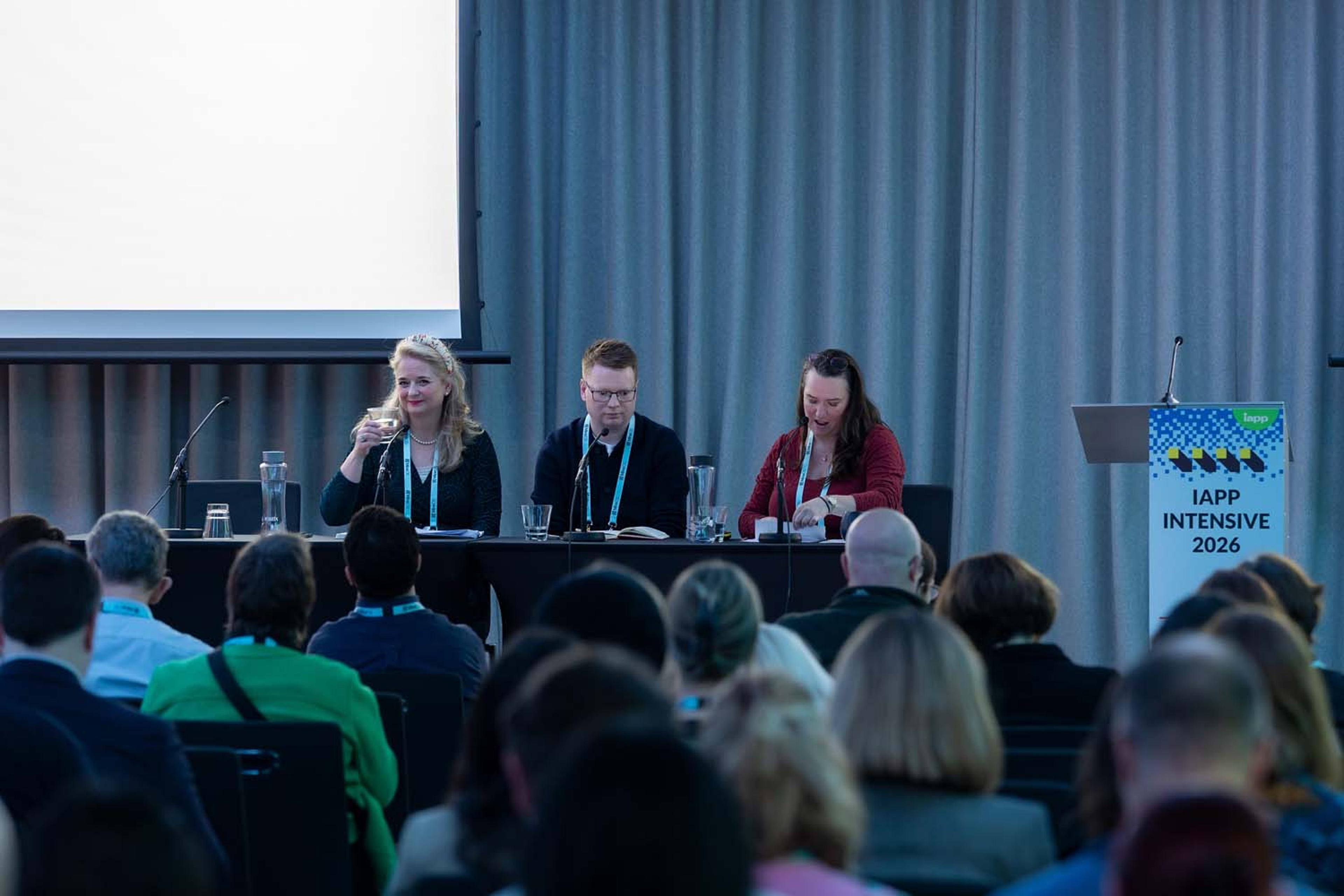 Panelists sit at a table onstage during a conference session while attendees watch from the audience. A podium with IAPP Intensive 2026 branding stands nearby.