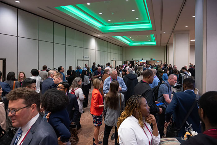 Crowded networking area with attendees standing and talking in groups. Bright overhead lighting and green accent lights illuminate the space.