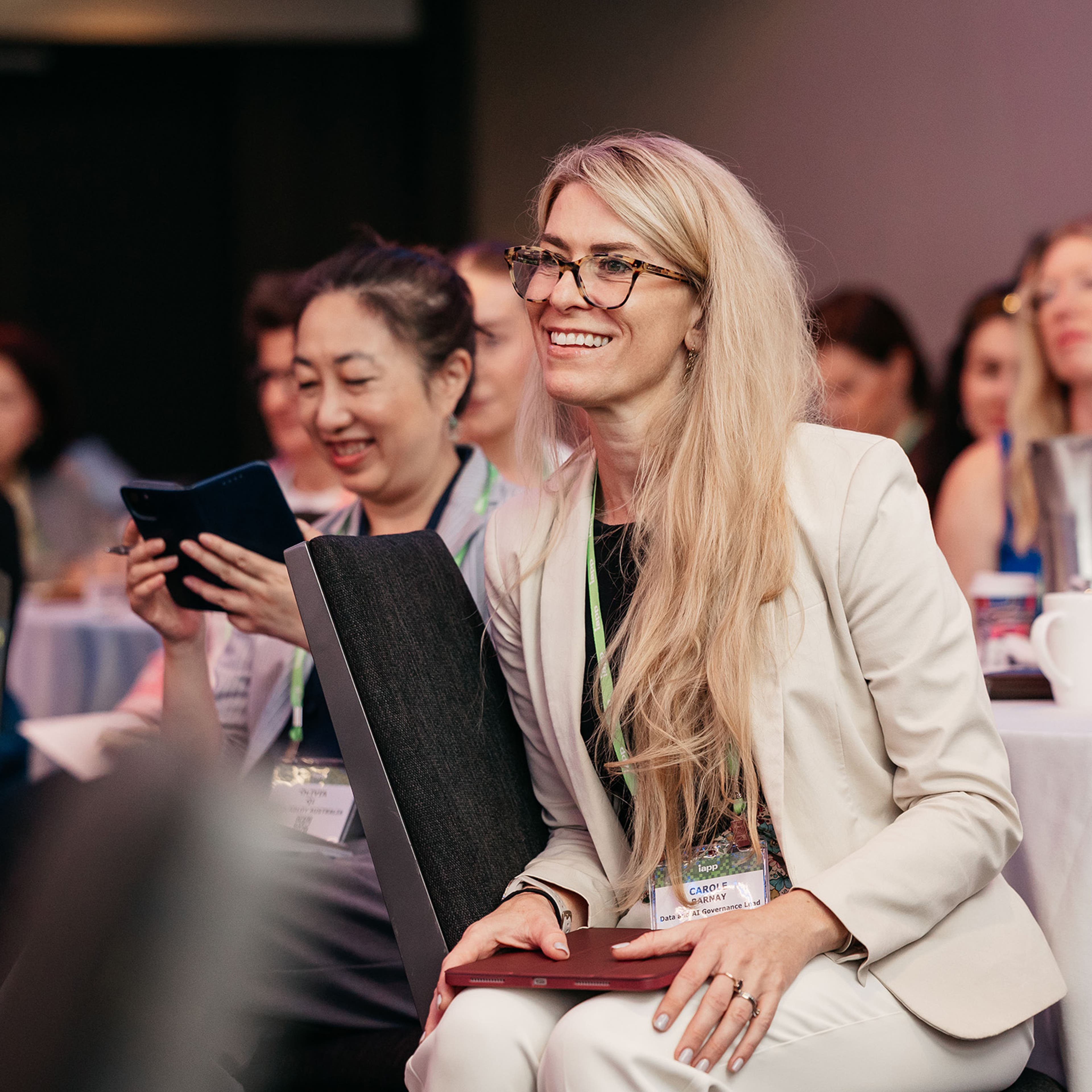 Attendee seated at a round table during a conference session, holding a tablet and listening to speakers.