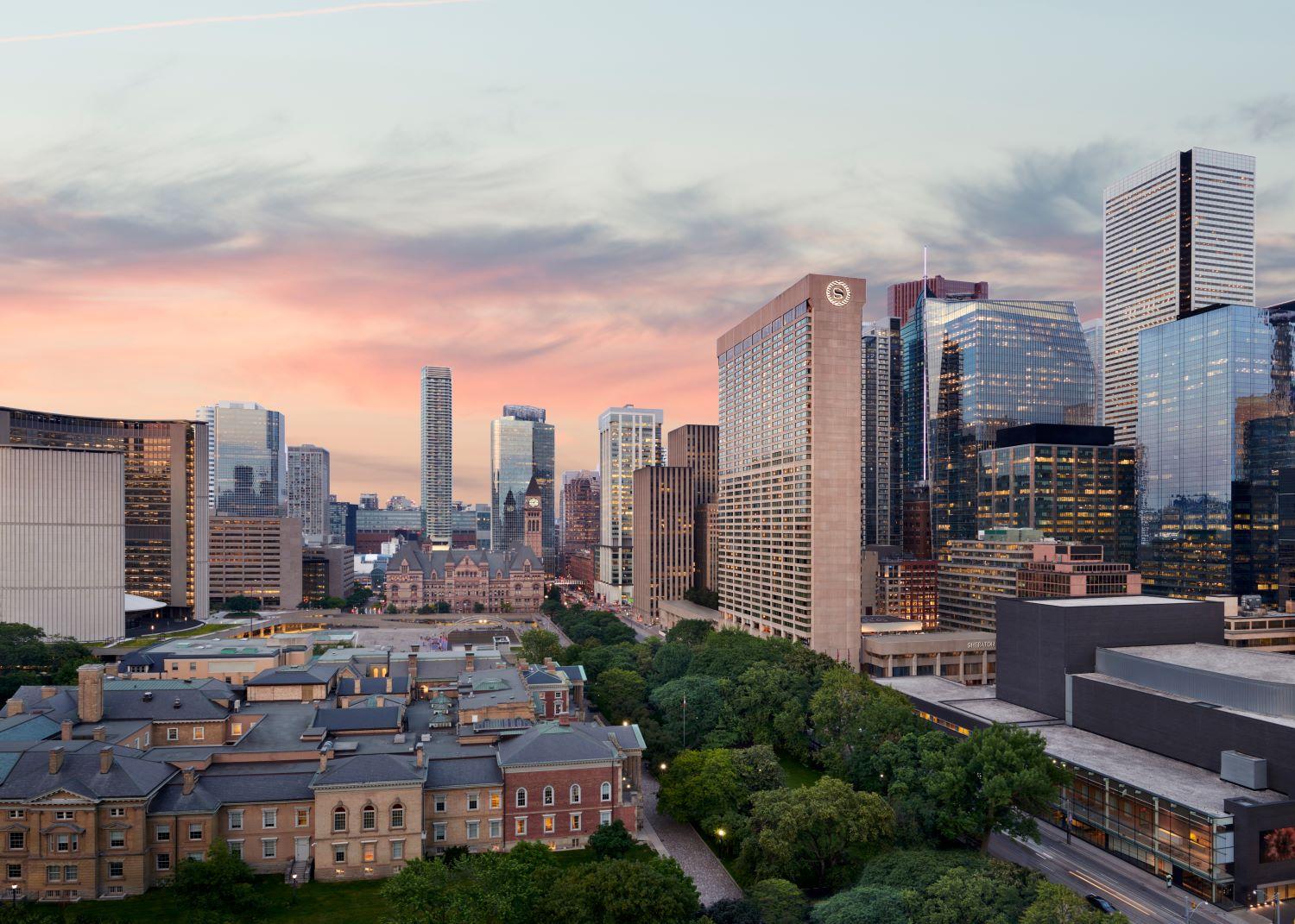 Cityscape of downtown Toronto at sunset, featuring the Sheraton Centre hotel prominently in the foreground. Surrounding the hotel are modern glass skyscrapers and historic buildings, with green trees and a park visible below. The sky is painted with soft pink and orange hues.