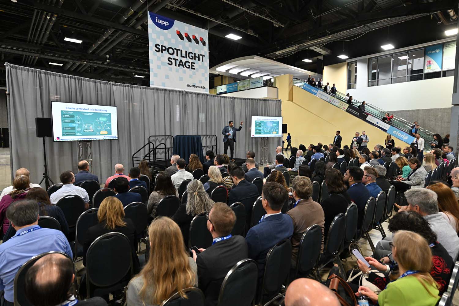 A presenter stands on the Spotlight Stage addressing a seated audience, flanked by presentation screens and framed by curtains and escalators in the background of the convention hall.