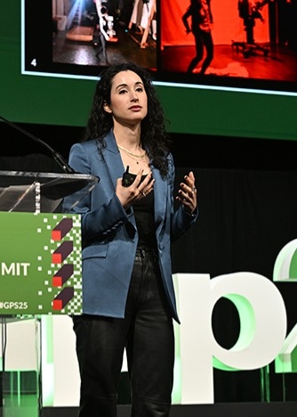 Presenter on stage wearing a blue blazer, gesturing with hands while speaking. Behind is a large screen displaying photos of people in a studio setting with bright lights and red backgrounds. The stage backdrop includes the event branding “GPS25.”