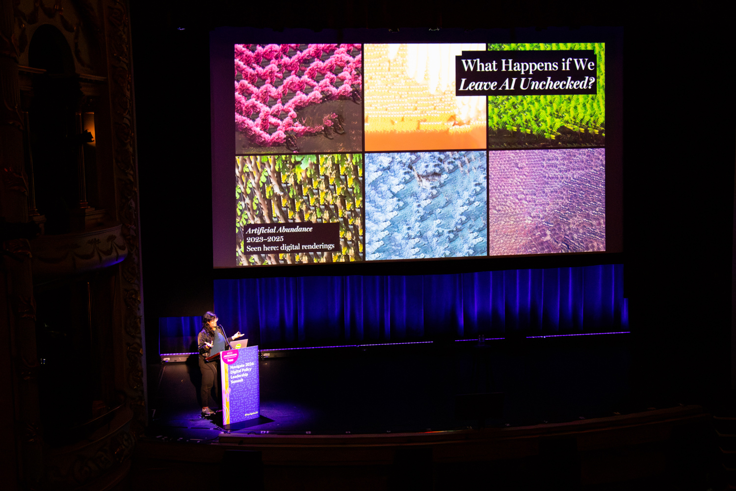 A speaker stands at a podium in a historic theater, delivering a presentation beneath a large screen displaying the slide title “What Happens if We Leave AI Unchecked?” alongside a colorful grid of abstract digital images.