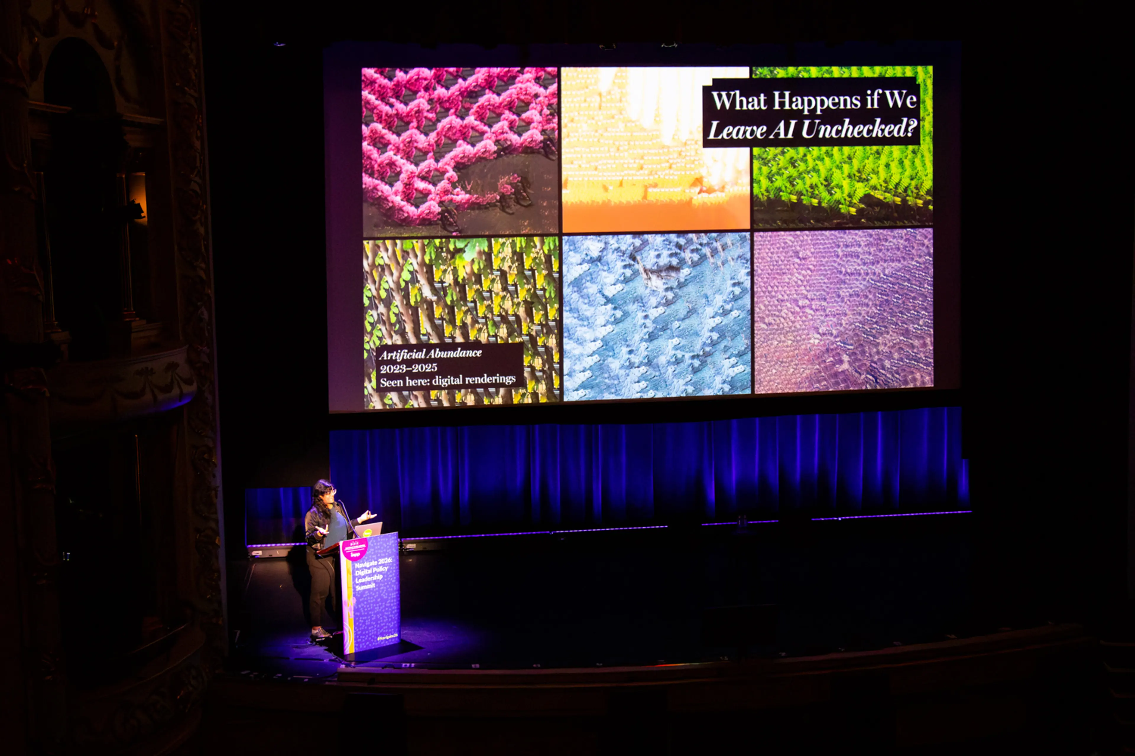 A speaker stands at a podium in a historic theater, delivering a presentation beneath a large screen displaying the slide title “What Happens if We Leave AI Unchecked?” alongside a colorful grid of abstract digital images.