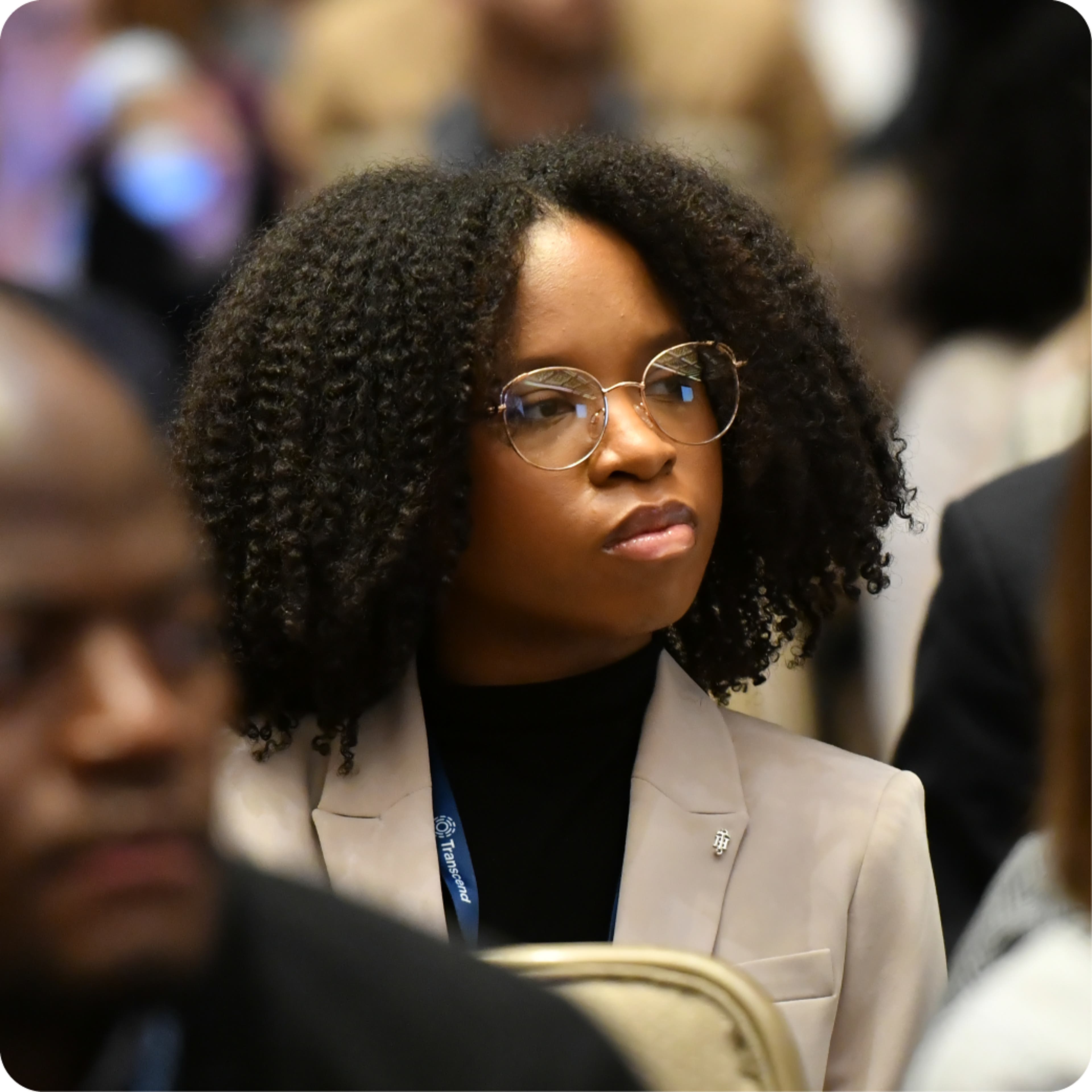 An african-american woman in glasses and a cream-colored jacket listening intently to a presentation