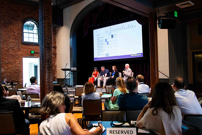 Conference session in a brick-walled venue with arched windows. A panel of four speakers sits on stage beneath a screen displaying a chart.