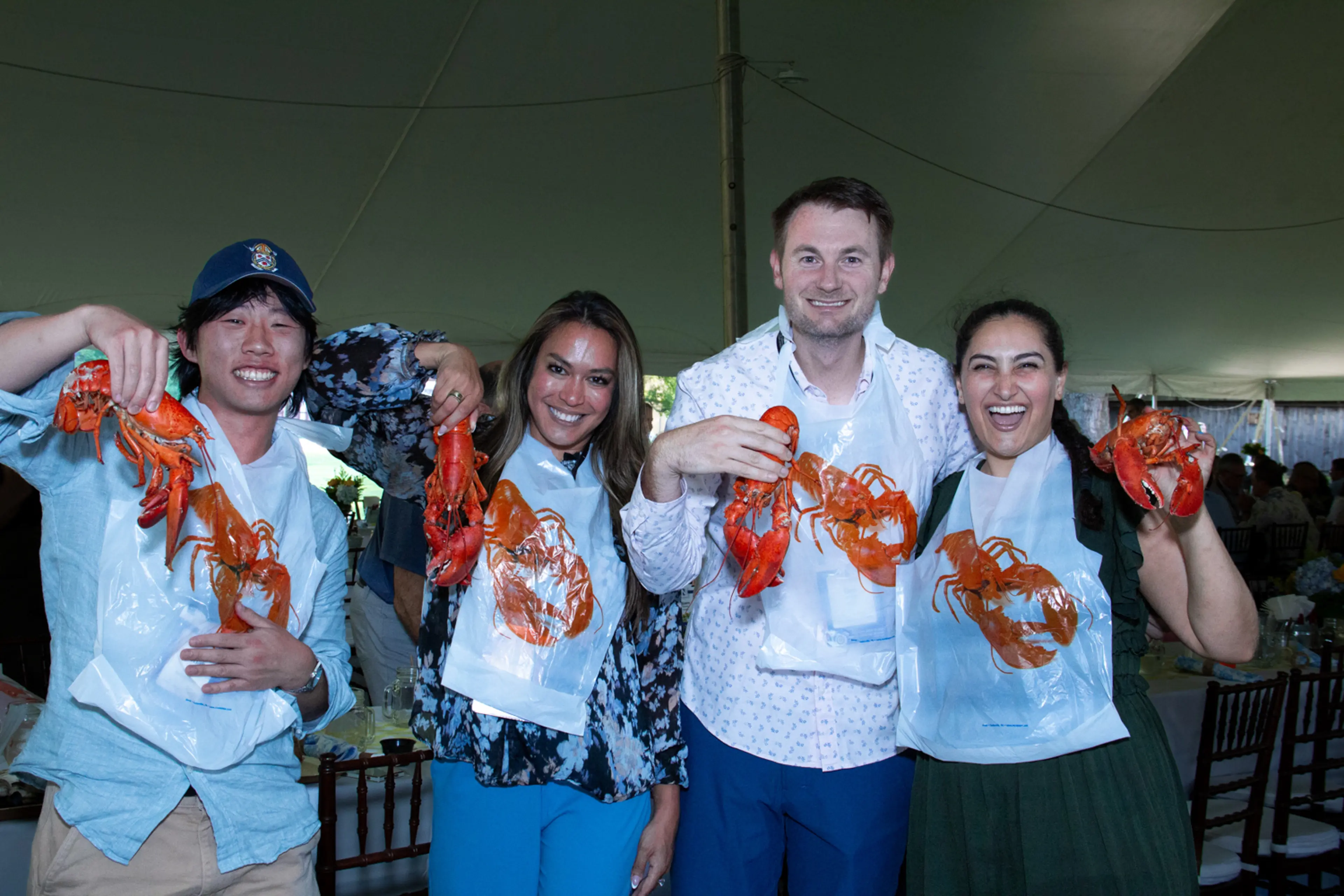 Four conference attendees wearing protective bibs pose together beneath a large event tent, each holding a bright red lobster during a lively networking gathering with banquet tables and guests visible in the background.