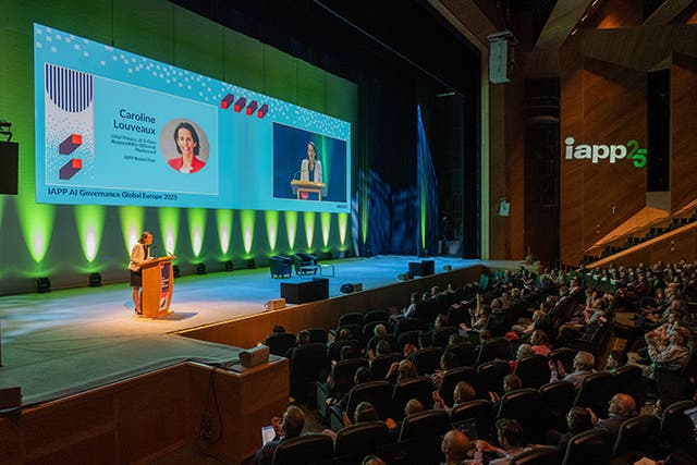 Large conference auditorium with rows of attendees seated facing a brightly lit stage. A speaker stands at a podium on the left side of the stage, which is illuminated with green and blue lighting. Two large screens display the speaker’s image and text reading “Caroline Louveaux” and “IAPP AI Governance Global Europe 2023.” The event branding “iapp25” is projected on the wooden wall to the right.