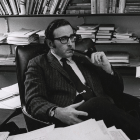 Black and white image of dr. alan westin sitting at a desk; he is surrounded by papers and is smoking a pipe.