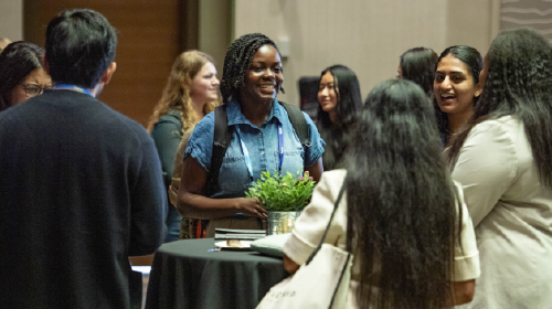 A young Black woman in a blue shirt stands by a small high-top table; other young people stand around her. They are all chatting.