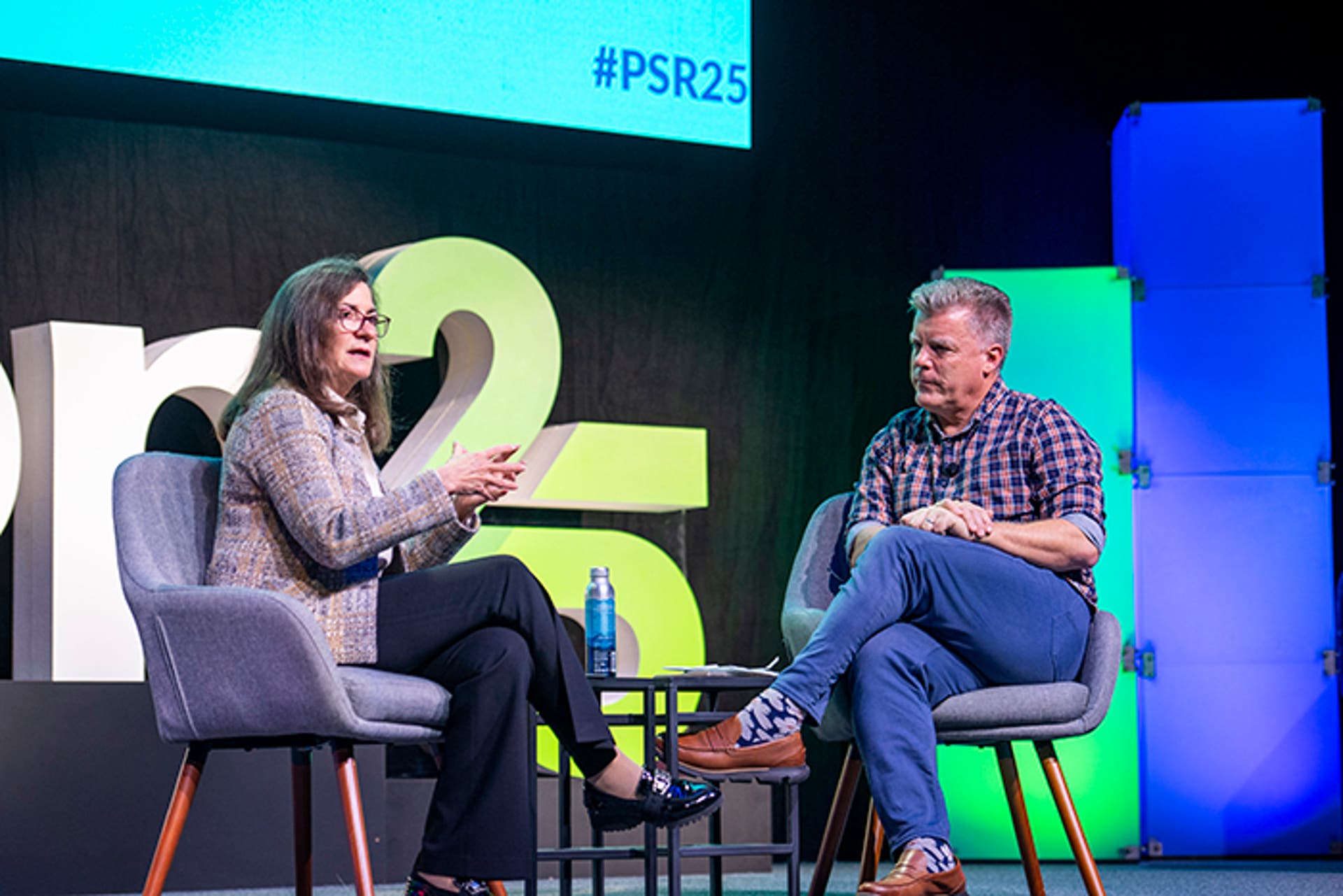 Two people seated on stage in gray chairs, engaged in a discussion. A small table with a water bottle is between them. Large “PSR25” signage and colorful light panels are in the background.