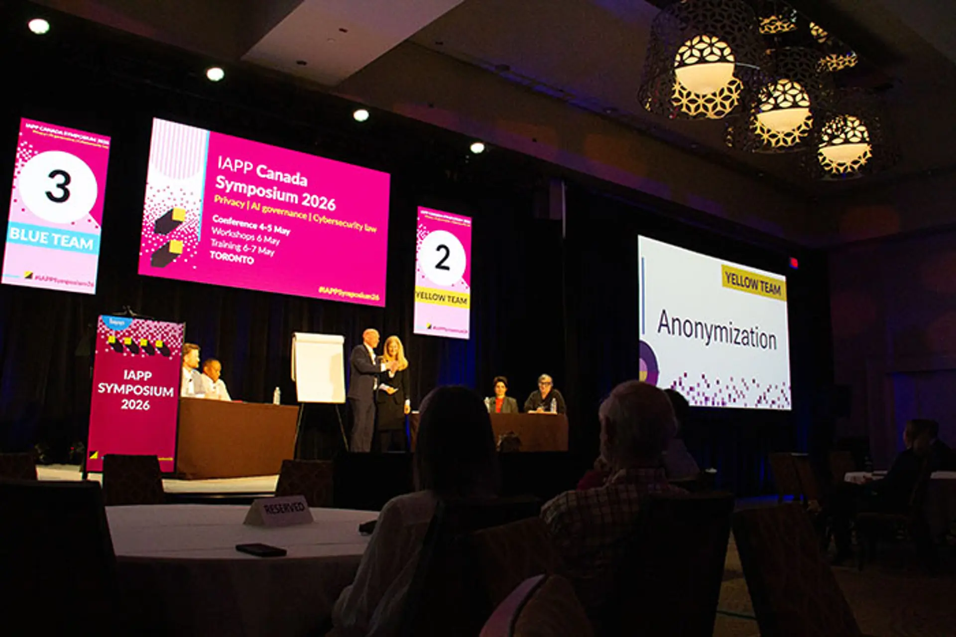 Wide view of a staged session at IAPP Canada Symposium 2026 featuring multiple speakers, large screens displaying event branding and “Anonymization,” and audience seated in a dimly lit conference hall.