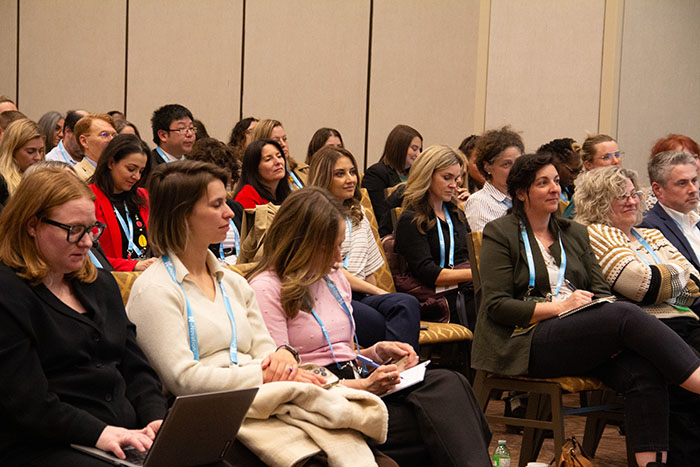 Audience seated in rows in a conference room, many wearing event lanyards and taking notes or using laptops while listening to a presentation.