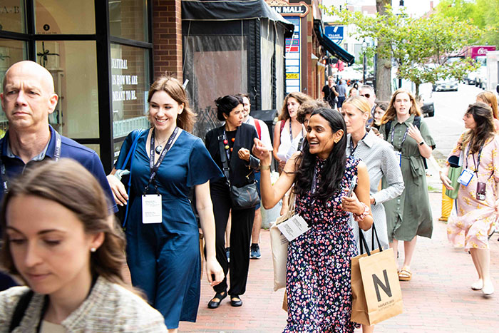 Group walking along a brick sidewalk in a downtown area, wearing conference badges and carrying tote bags. Storefronts and trees line the street.