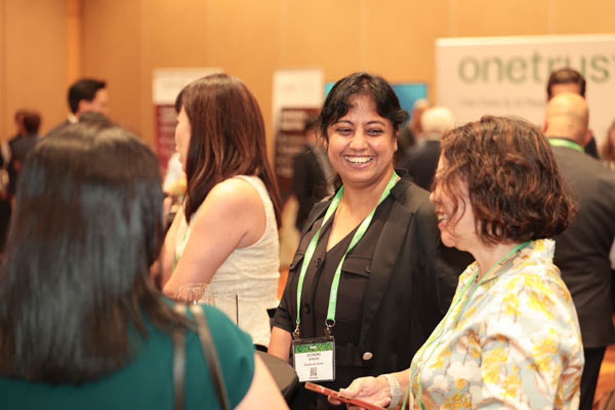 Large conference room with attendees seated at round tables facing a stage. Screens display “Welcome and Introductions” and speaker details.