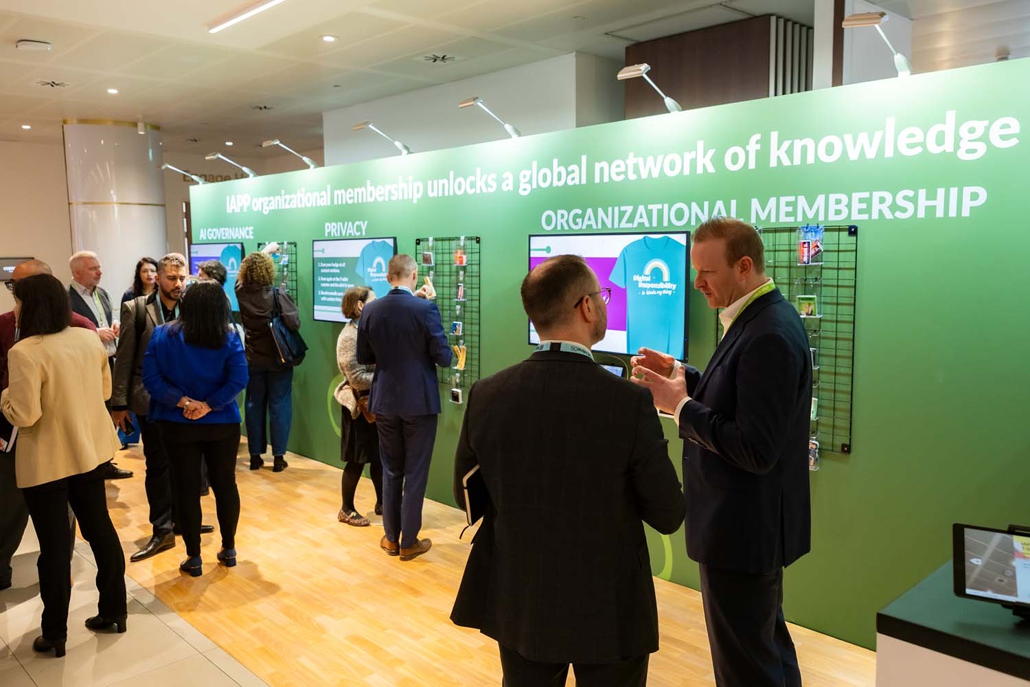 Conference attendees gather around an IAPP organizational membership display highlighting privacy and governance themes. Digital screens and informational panels line a green branded wall.
