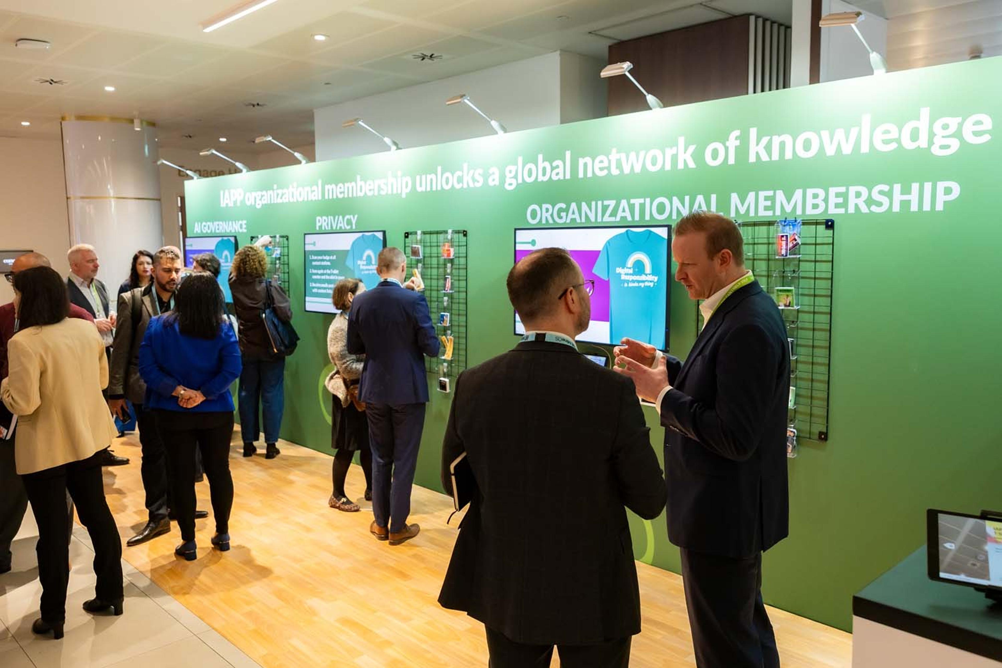 Conference attendees gather around an IAPP organizational membership display highlighting privacy and governance themes. Digital screens and informational panels line a green branded wall.