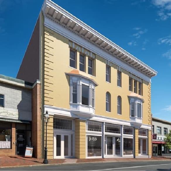 Historic three-story building with a pale yellow facade and white trim, featuring large bay windows on the second floor and tall rectangular windows above. The ground level has multiple glass storefronts, and the building is set along a brick sidewalk under a clear blue sky.