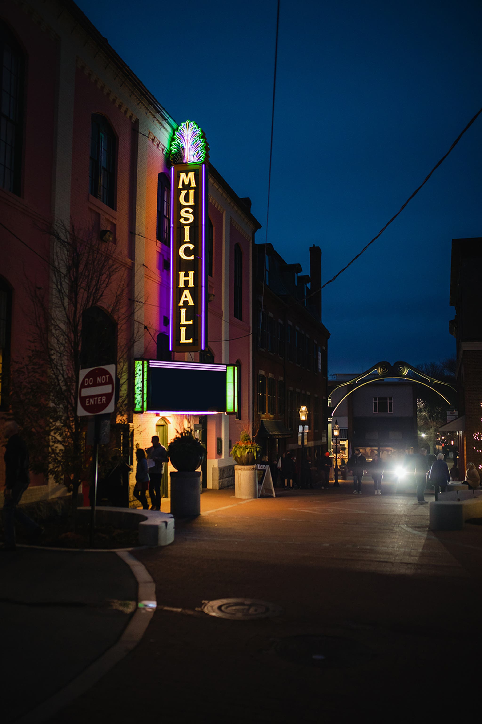 Nighttime street scene featuring the Music Hall building with a tall vertical neon sign glowing green and purple that reads “MUSIC HALL.” The marquee below is illuminated, and the surrounding area includes brick buildings, potted plants, and a few pedestrians. A car with headlights on is visible in the distance under a dark blue sky.