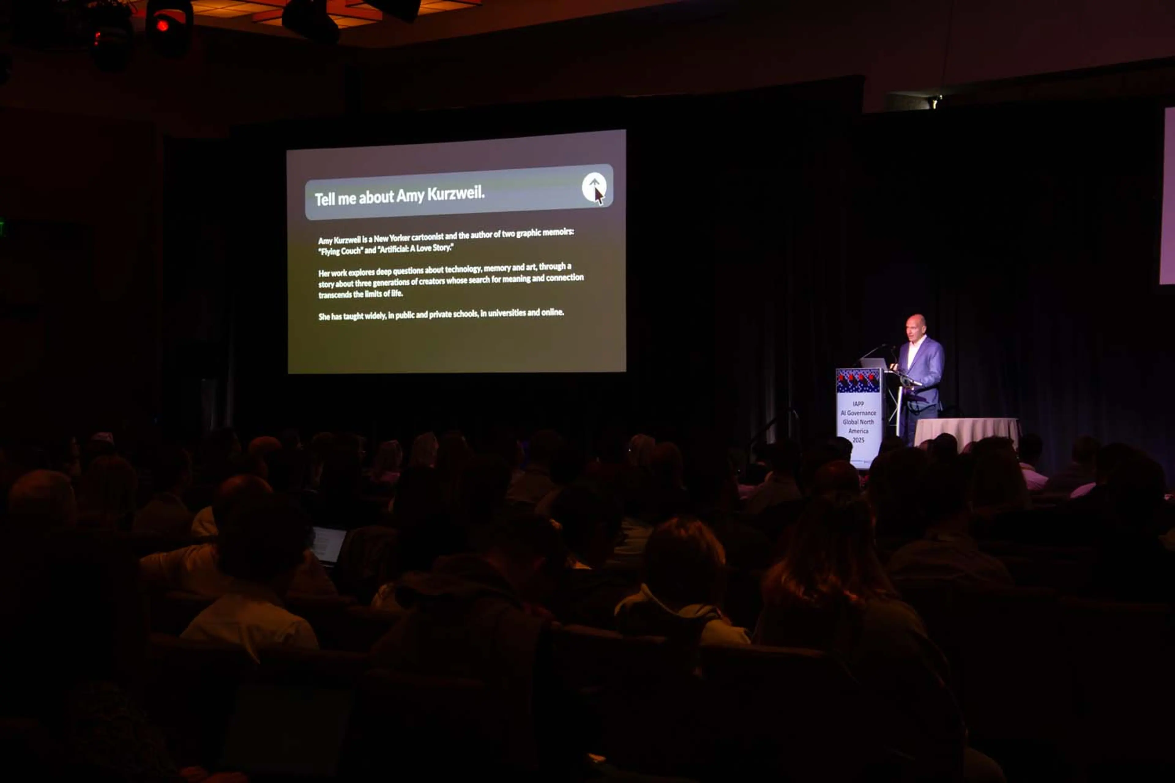 A presenter stands at a podium on a dimly lit stage while a large screen behind displays a slide titled “Tell me about Amy Kurzweil,” with the audience silhouetted in the foreground.