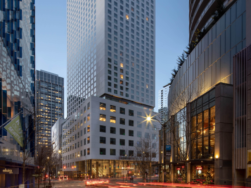 A tall, angular tower with a grid of windows stands against a clear evening sky, framed by modern buildings and a lively downtown street illuminated by headlights and storefront lights.