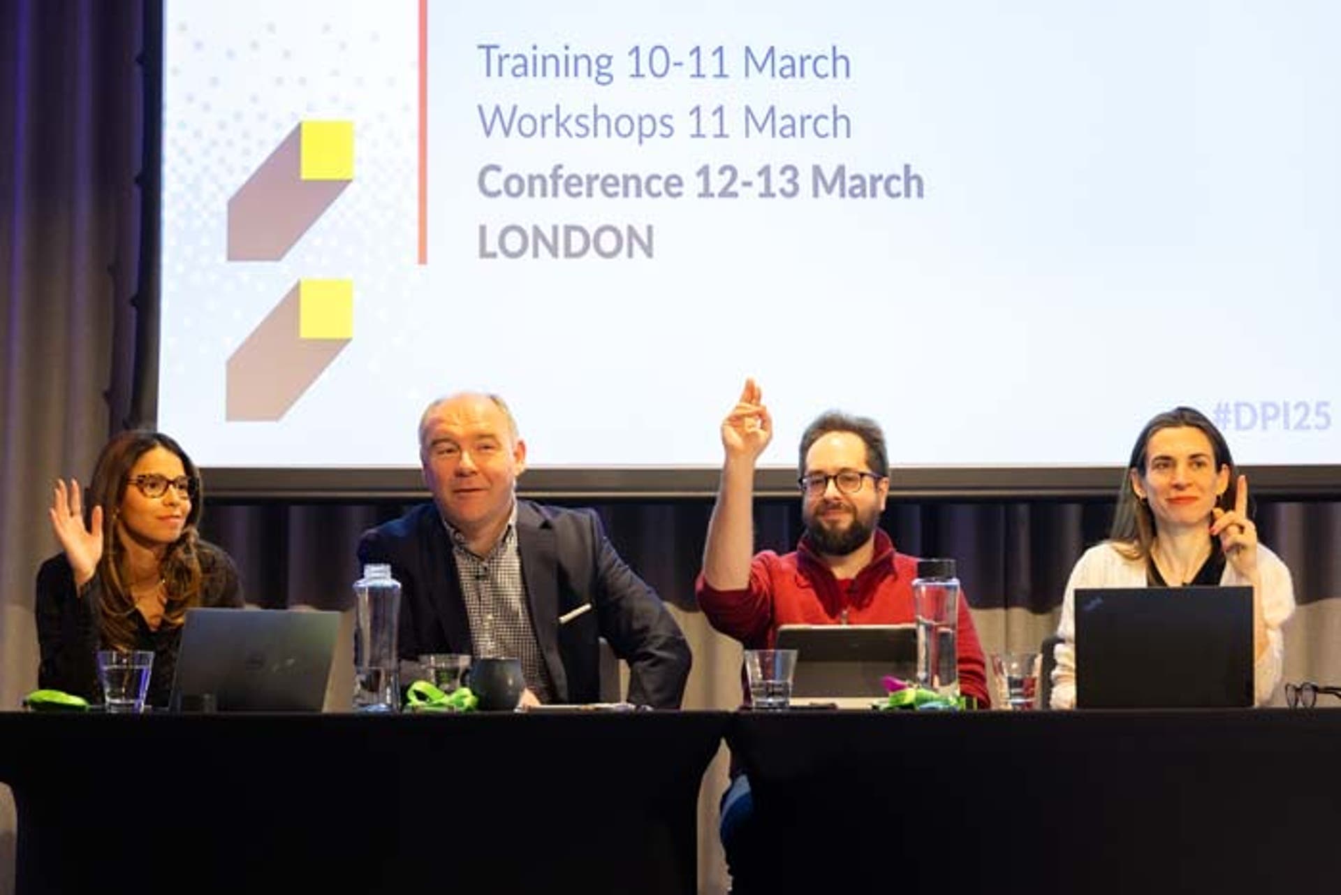 Two presenters at a conference. On the right is a woman who is raising her hand to make a point.