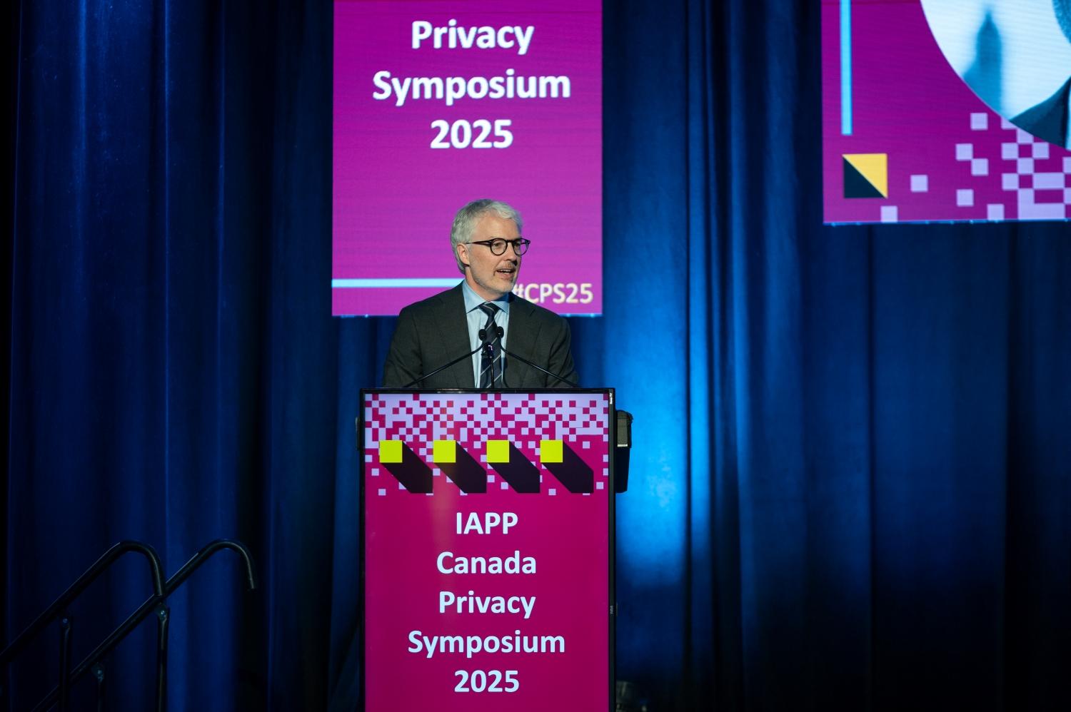 Speaker standing at a podium with a bright pink sign that reads “IAPP Canada Privacy Symposium 2025.” Behind the podium is a large blue curtain backdrop, and a matching pink screen above displays the text “Privacy Symposium 2025.”