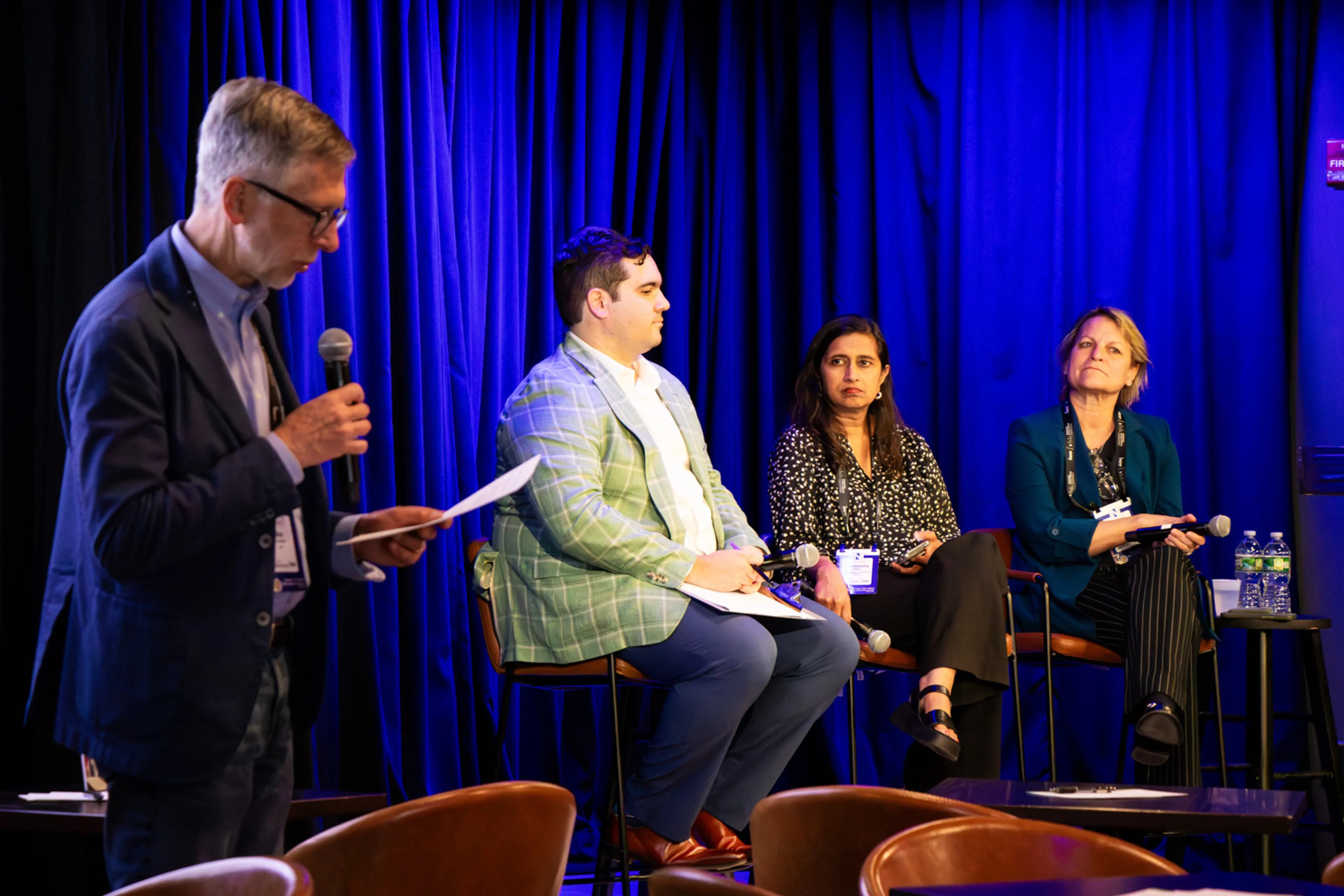 A moderator stands at the side of a stage holding a microphone and notes while three panelists sit in discussion against a backdrop of deep blue curtains, with microphones, water bottles, and audience seating visible in the foreground.