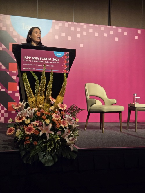 A speaker addresses attendees from a podium at the IAPP Asia Forum 2026. A large floral arrangement is positioned in front of the podium, and a pink conference backdrop and stage seating are visible in the background.