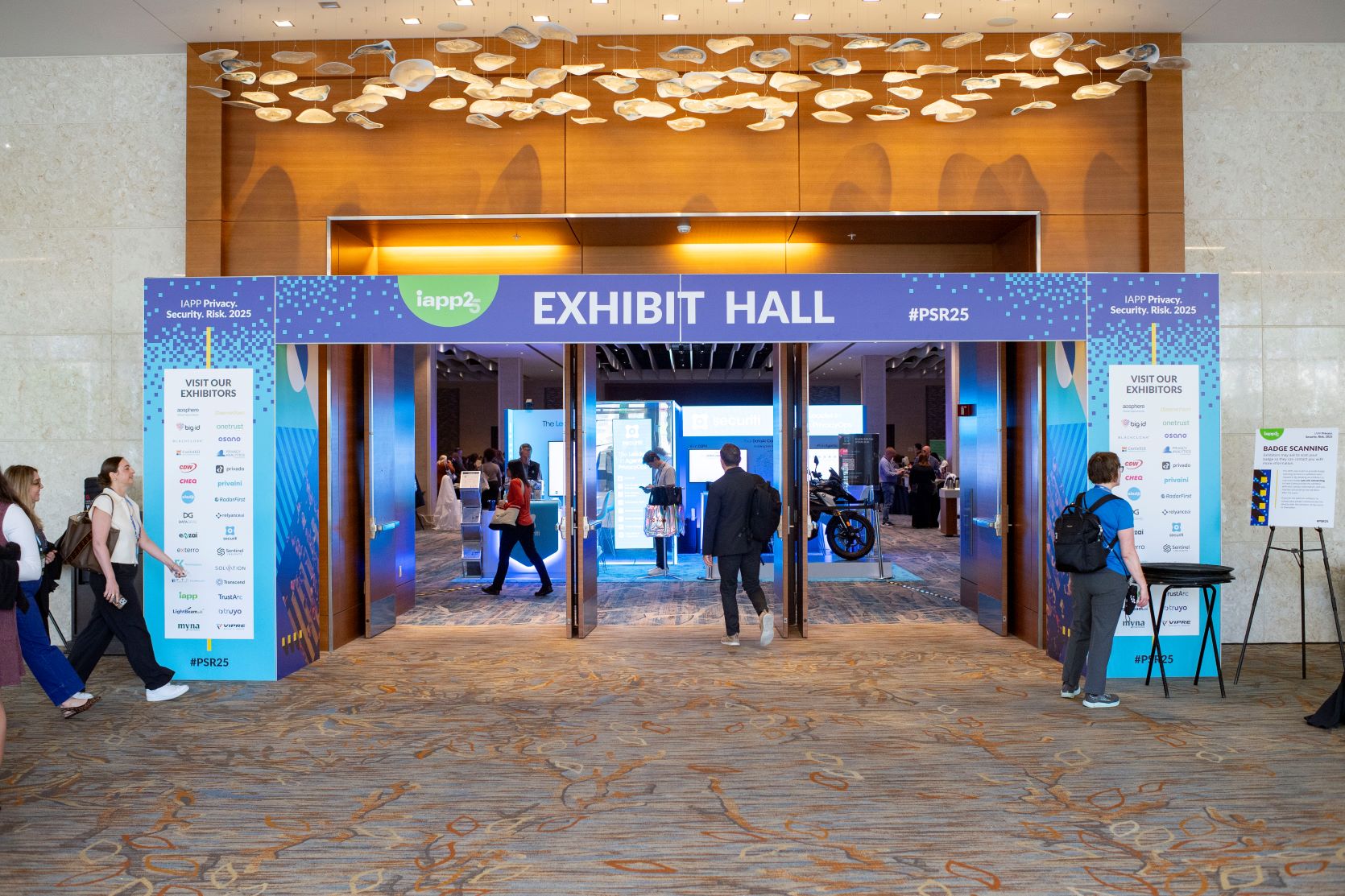 Entrance to an exhibit hall at a conference, framed by a large blue banner reading “EXHIBIT HALL” with event branding “IAPP Privacy. Security. Risk. 2025” and hashtag #PSR25. The banner lists “Visit Our Exhibitors” on both sides. Inside the hall, bright display booths and screens are visible, and several attendees are walking in and out of the entrance. The space features patterned carpet and modern ceiling lights.