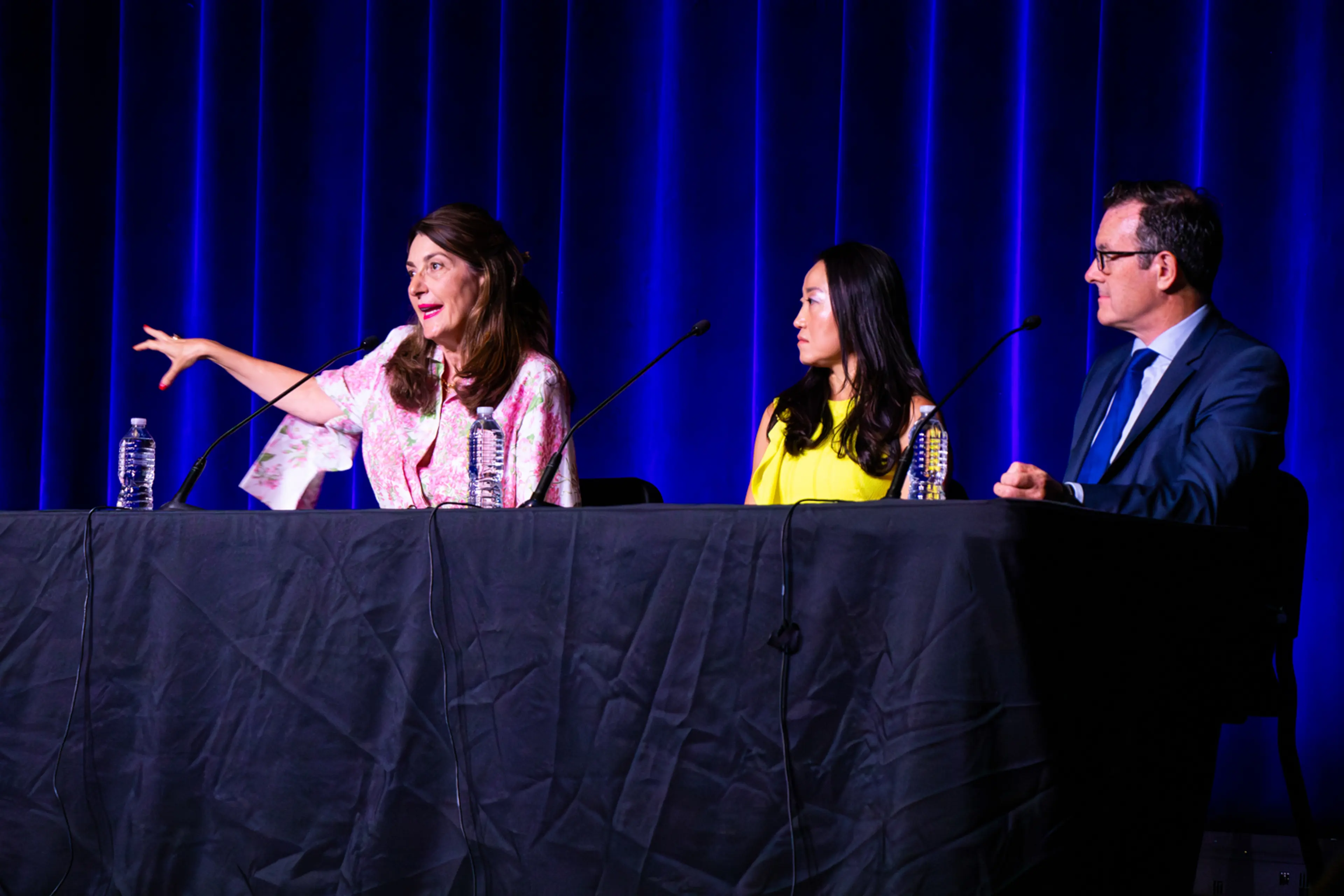 Three panelists sit behind a long table on stage during a conference discussion, speaking into microphones against a backdrop of illuminated blue curtains, while water bottles and presentation equipment are arranged across the table.