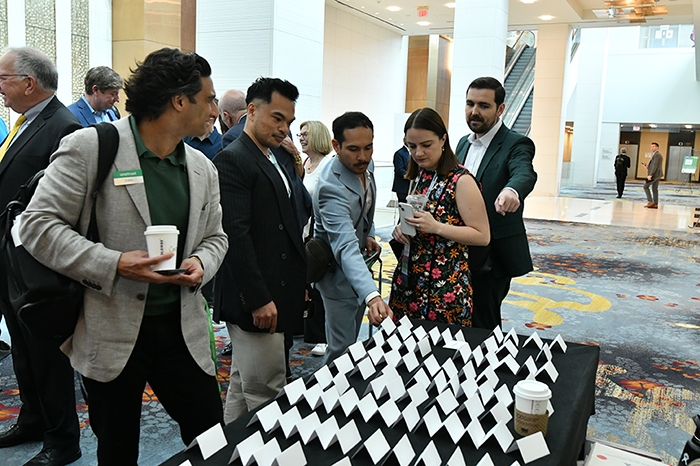 Group of attendees gathered around a registration table covered with rows of white name badges. Several people are holding coffee cups, and the background shows a spacious lobby with patterned carpet and escalators.